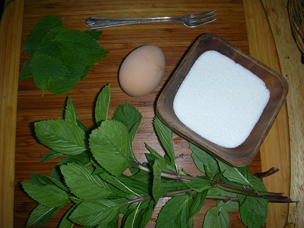 Fresh peppermint leaves, an egg, white sugar, and a fork on a wooden cutting board